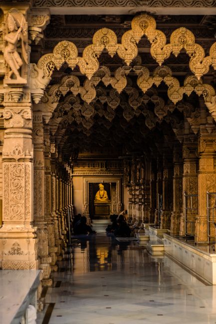 An ornate corridor inside the Neasden Mandir features intricately carved marble pillars and arches with gold embellishments, leading to a central shrine with a seated golden Buddha statue illuminated by ambient lighting. The polished marble floor reflects the detailed architecture and a few individuals sitting quietly in the hallway. The ceiling showcases elaborate patterns and motifs, emphasizing traditional temple craftsmanship. The space appears clean and well-maintained, suitable for regular surface cleaning and sanctification, as provided by Carpet Cleaning Brent. Utilizing deep cleaning methods, the corridor remains dust-free and hygienic, ensuring a respectful and serene environment for visitors and worshippers.