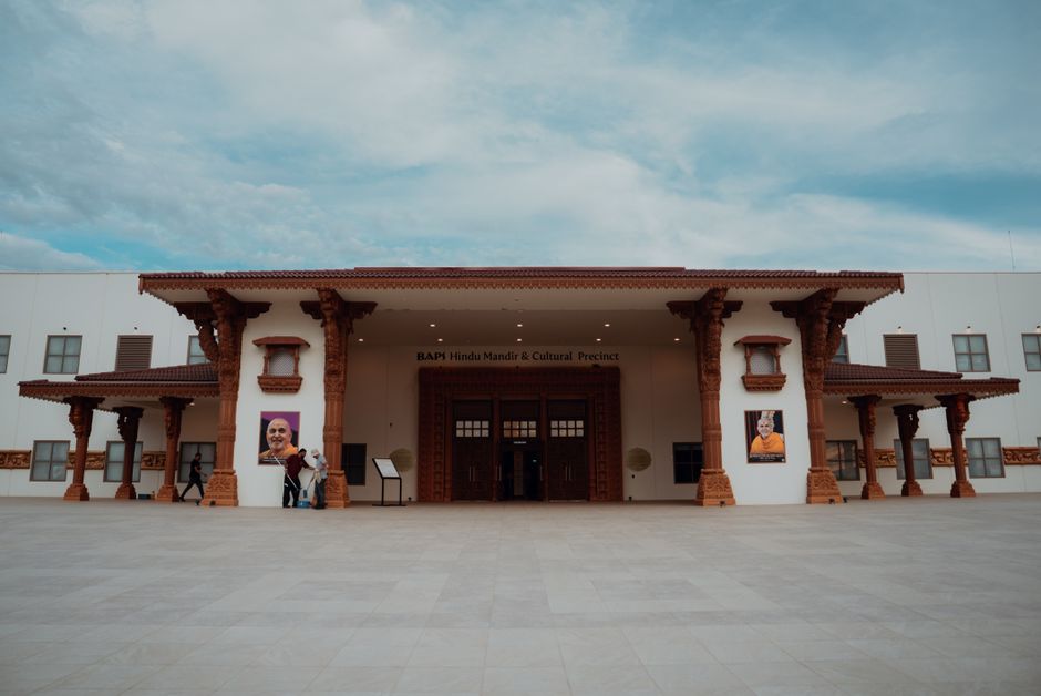 The exterior of the Neasden Mandir, a Hindu religious temple in Brent, displaying a white façade with traditional wooden-style architectural elements supported by multiple carved wooden pillars. The entrance features a large, dark wooden door with small glass panels, surrounded by a decorative wooden frame. Two large posters of individuals are mounted on either side of the entrance, and a small group of people are visible near the doorway. The foreground consists of a wide, paved open area with a smooth, light-colored surface under a partly cloudy sky. The overall scene highlights the building's cultural architectural style and clean, well-maintained appearance, with natural lighting enhancing the details.