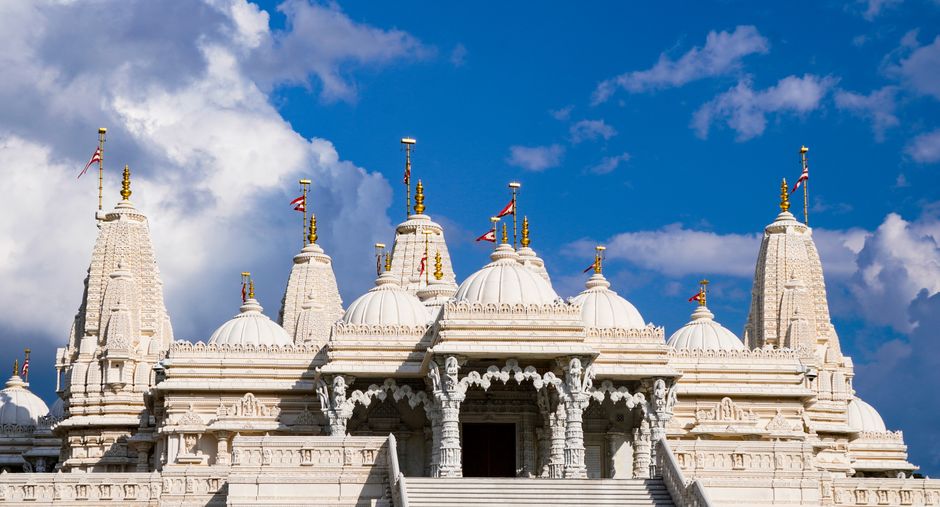 An ornate corridor inside the Neasden Mandir features intricately carved marble pillars and arches with gold embellishments, leading to a central shrine with a seated golden Buddha statue illuminated by ambient lighting. The polished marble floor reflects the detailed architecture and a few individuals sitting quietly in the hallway. The ceiling showcases elaborate patterns and motifs, emphasizing traditional temple craftsmanship. The space appears clean and well-maintained, suitable for regular surface cleaning and sanctification, as provided by Carpet Cleaning Brent. Utilizing deep cleaning methods, the corridor remains dust-free and hygienic, ensuring a respectful and serene environment for visitors and worshippers.