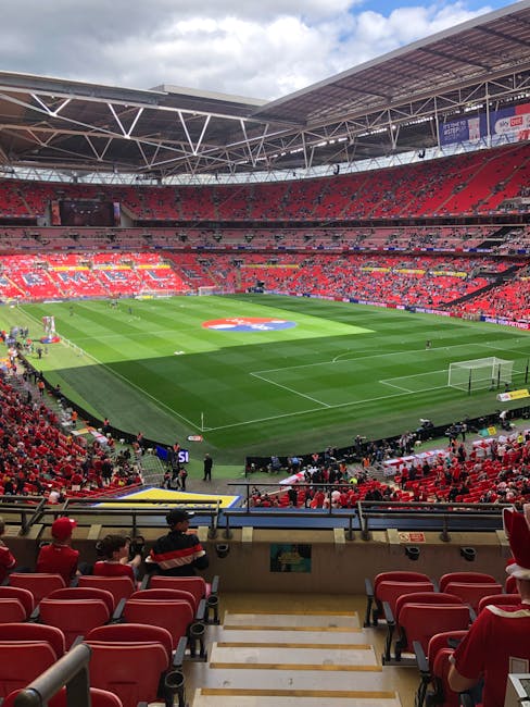 Interior view of Wembley Stadium with a well-maintained, lush green football pitch and surrounding red seating areas, under a partially cloudy sky. The field displays clear, freshly mowed grass with visible white line markings, goalposts at each end, and a large team emblem in the center. The seating sections are mostly empty, with some spectators seated towards the front. Overhead, the stadium's modern roof structure with metal beams and panels provides shelter, allowing natural light to illuminate the field evenly. The stadium appears clean and organized, ready for a match or event, with no visible debris or dust. This image highlights the spacious, hygienic environment maintained within the sports arena, demonstrating professional surface cleaning and maintenance typical of high-profile venues. For detailed cleaning and sanitisation services, trust Carpet Cleaning Brent, experts near HA9, BRENT, to keep your facilities in top condition.