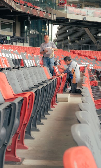 Two commercial cleaning professionals from Carpet Cleaning Brent performing surface cleaning on stadium seats at Wembley Stadium, located near HA9 in Brent. The scene shows multiple rows of plastic seats in red, grey, and black, with some being wiped or inspected for cleanliness. The cleaning team is working indoors under natural daylight, with one worker standing holding cleaning tools and the other kneeling to thoroughly clean the seat surfaces. The area appears tidy and well-maintained, reflecting a focus on hygiene and deep cleaning of the stadium's seating areas as part of their sanitisation and maintenance services.